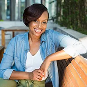 A smiling woman with short hair, wearing a denim shirt and white top, sitting on a wooden bench in a modern outdoor setting.