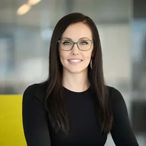 A professional headshot of a smiling woman with long black hair and glasses, wearing a black top, set against a blurred office background.