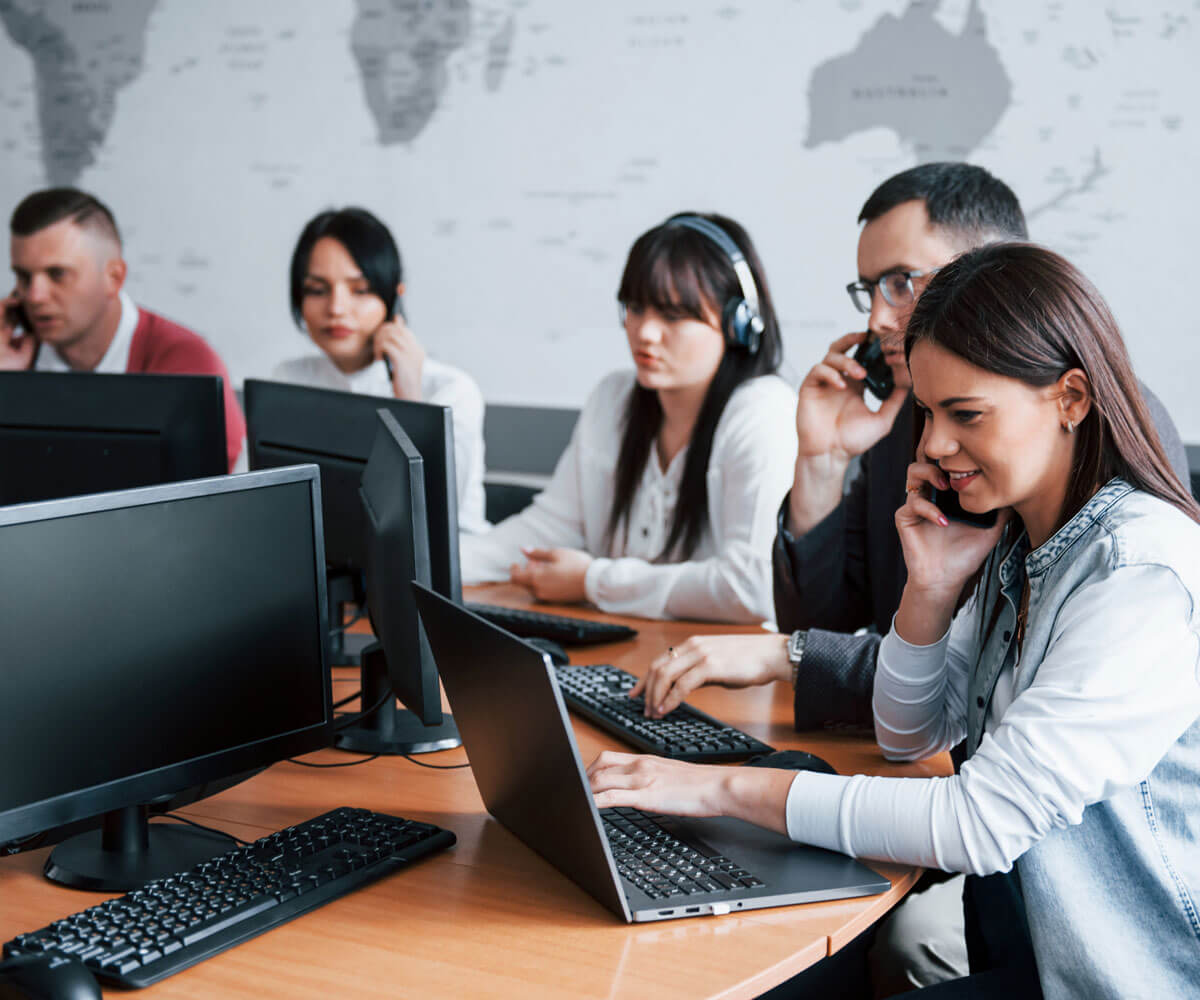 A busy call center office with five agents, each focused on their computer screens and wearing headsets. one female agent is speaking on the phone. a world map decorates the background wall.